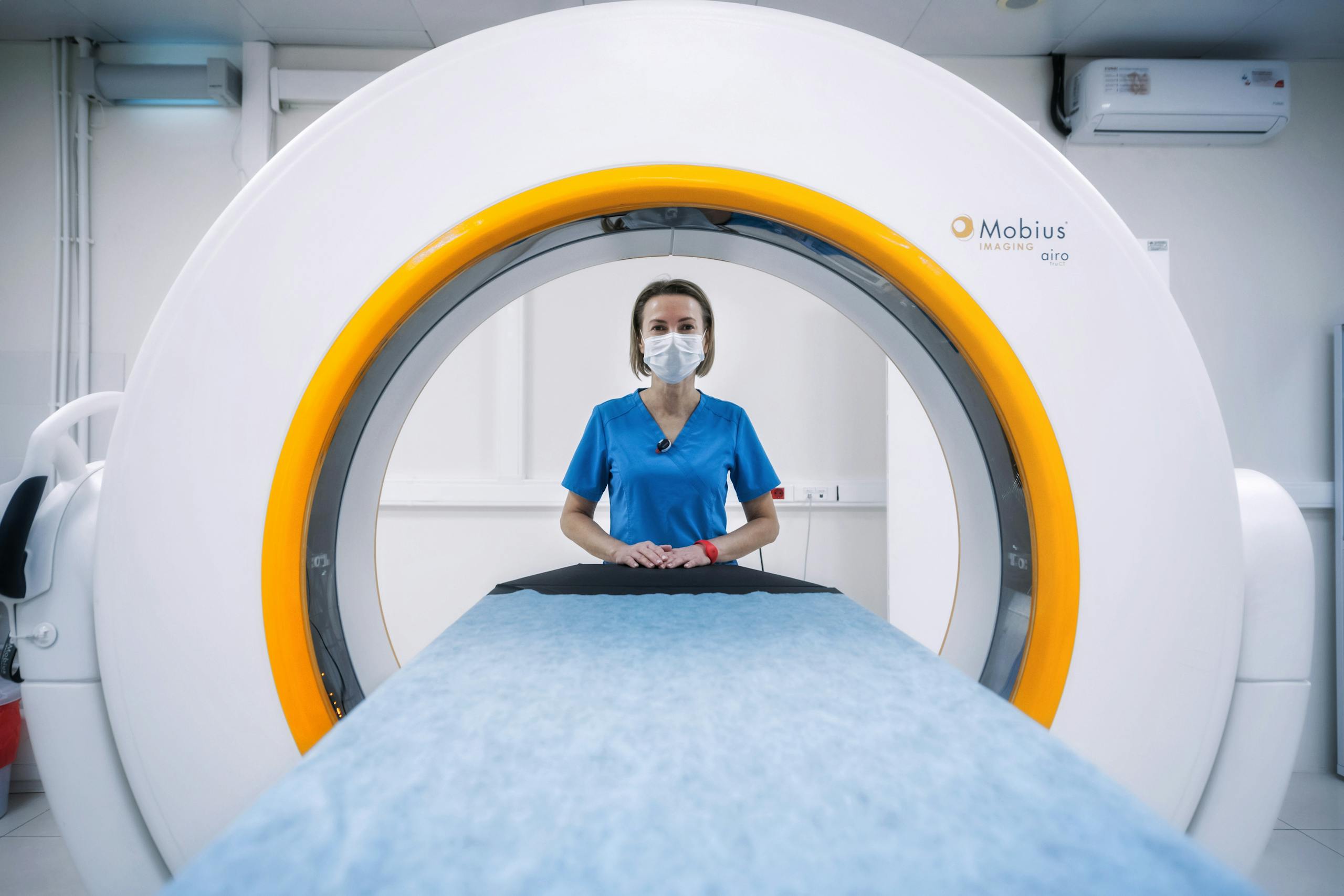 Nurse in blue scrubs and mask stands by an MRI machine, ready for a scan.